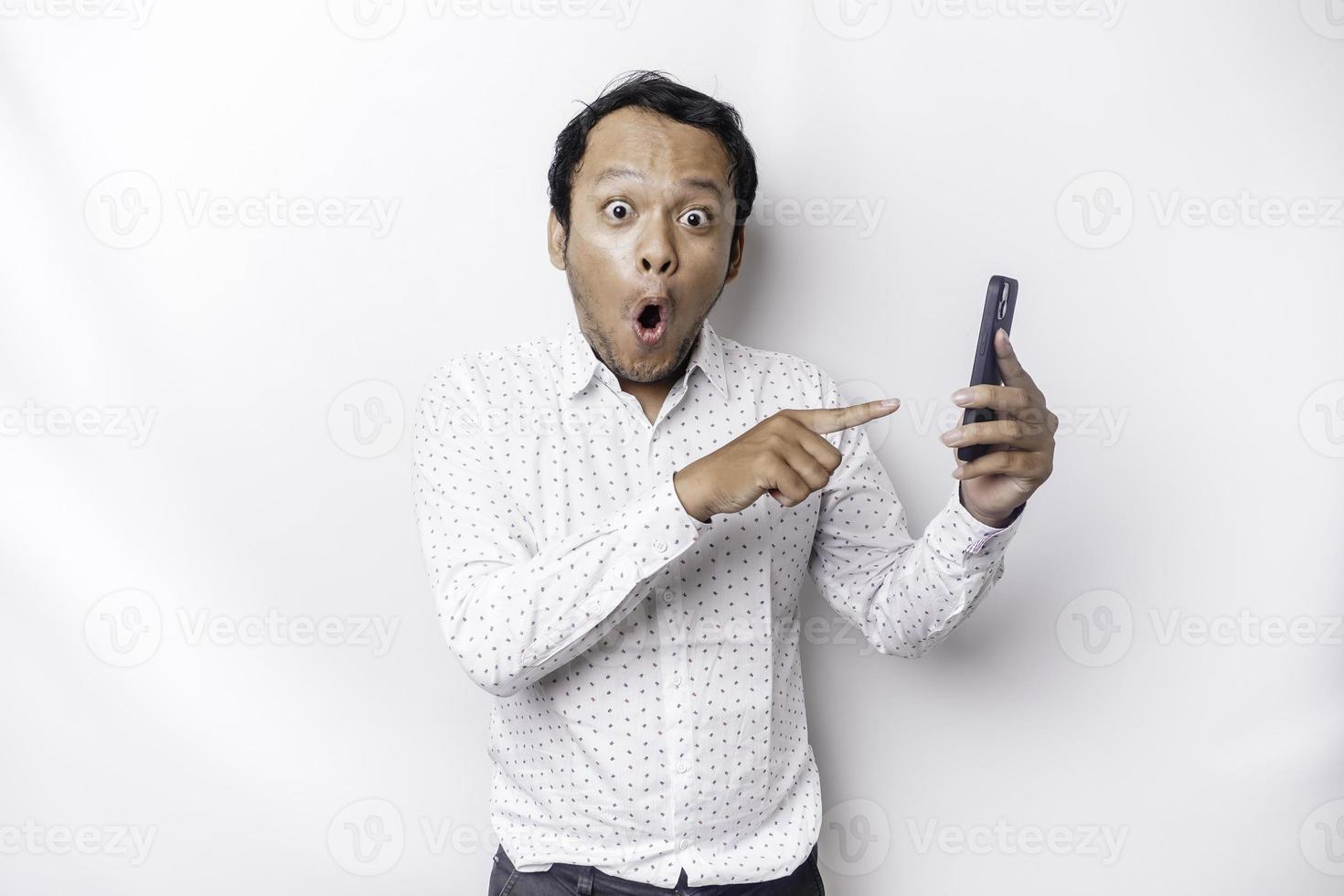 chocado ásia homem vestindo uma branco camisa e segurando dele telefone, isolado de branco fundo foto