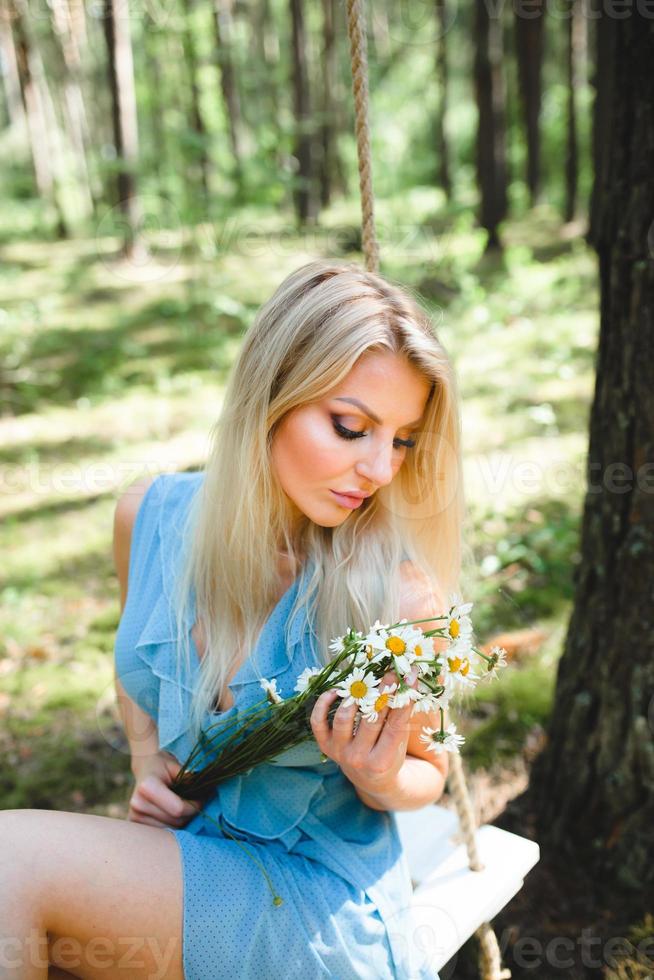jovem lindo mulher dentro curto azul vestir posando em uma corda balanço com ramalhete do branco flores foto