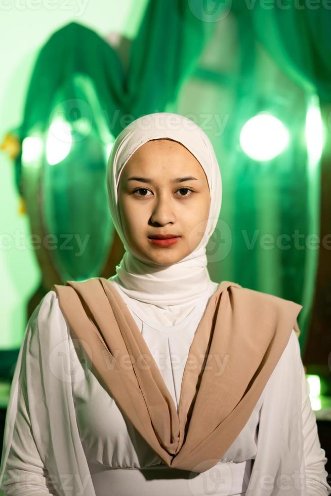 uma muçulmano mulher com uma branco lenço de cabeça e branco roupas é sentado dentro a meio do uma verde quarto sem Maquiagem foto