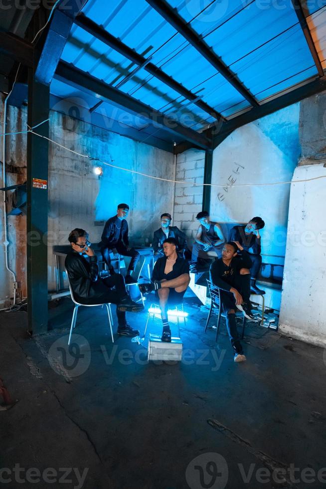 uma grupo do ásia adolescentes dentro Preto roupas sentado juntos dentro uma Preto construção com uma azul luz em a quarto foto