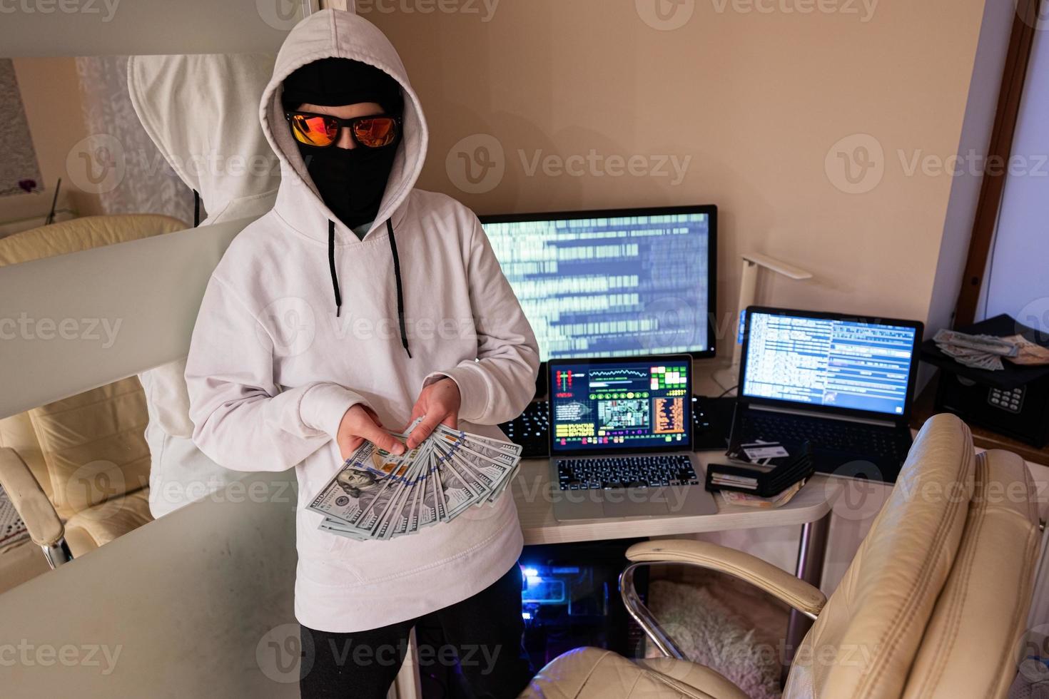 Garoto hacker com dinheiro dólares. Internet roubo . homem vestindo uma balaclava atrás uma computador portátil. foto