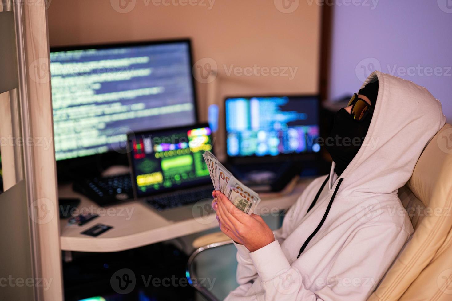 Garoto hacker com dinheiro dólares. Internet roubo . homem vestindo uma balaclava Sentou atrás uma computador portátil. foto