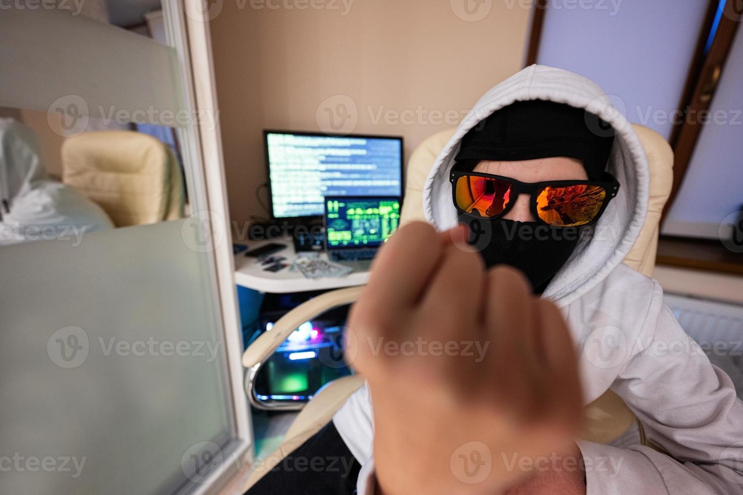 Garoto hacker fraudulentamente usar crédito cartão para pagamento. Internet roubo mostra punho . homem vestindo uma balaclava e segurando uma crédito cartão enquanto Sentou atrás uma computador portátil. foto