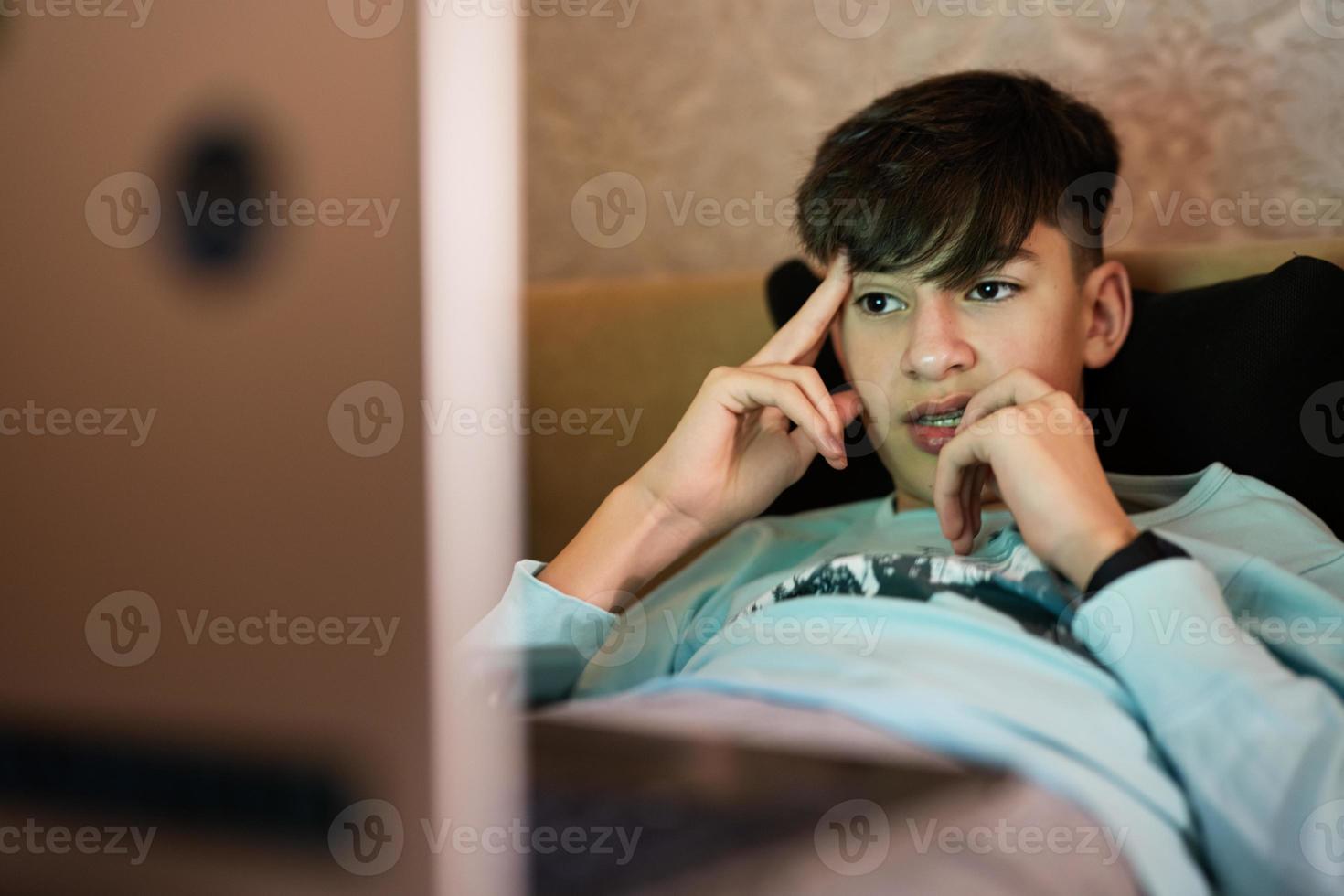 jovem adolescente Garoto dentro frente do uma computador portátil em uma cama às tarde. foto