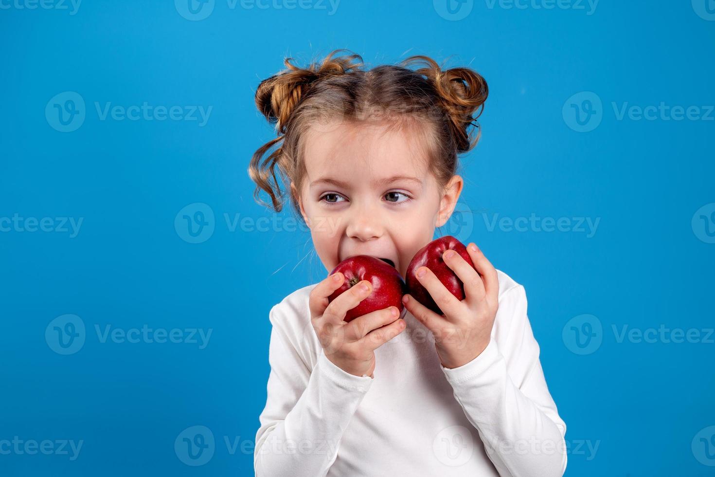 fofa pequeno menina com cachos dentro uma listrado vestir é segurando uma grande vermelho maçã dentro dela mãos. azul fundo. útil produtos para crianças. saudável lanche. espaço para texto. Alto qualidade foto