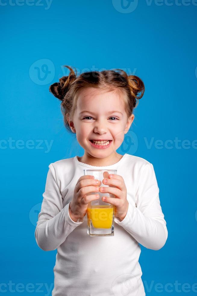 pequeno menina com uma engraçado Penteado dentro uma branco camiseta bebidas suco a partir de uma vidro. azul fundo. útil produtos para crianças. espaço para texto. Alto qualidade foto