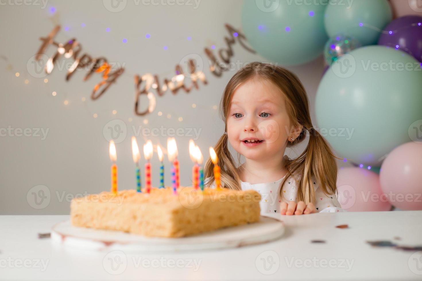 fofa pequeno menina golpes Fora velas em uma aniversário bolo às casa contra uma pano de fundo do balões. criança aniversário foto