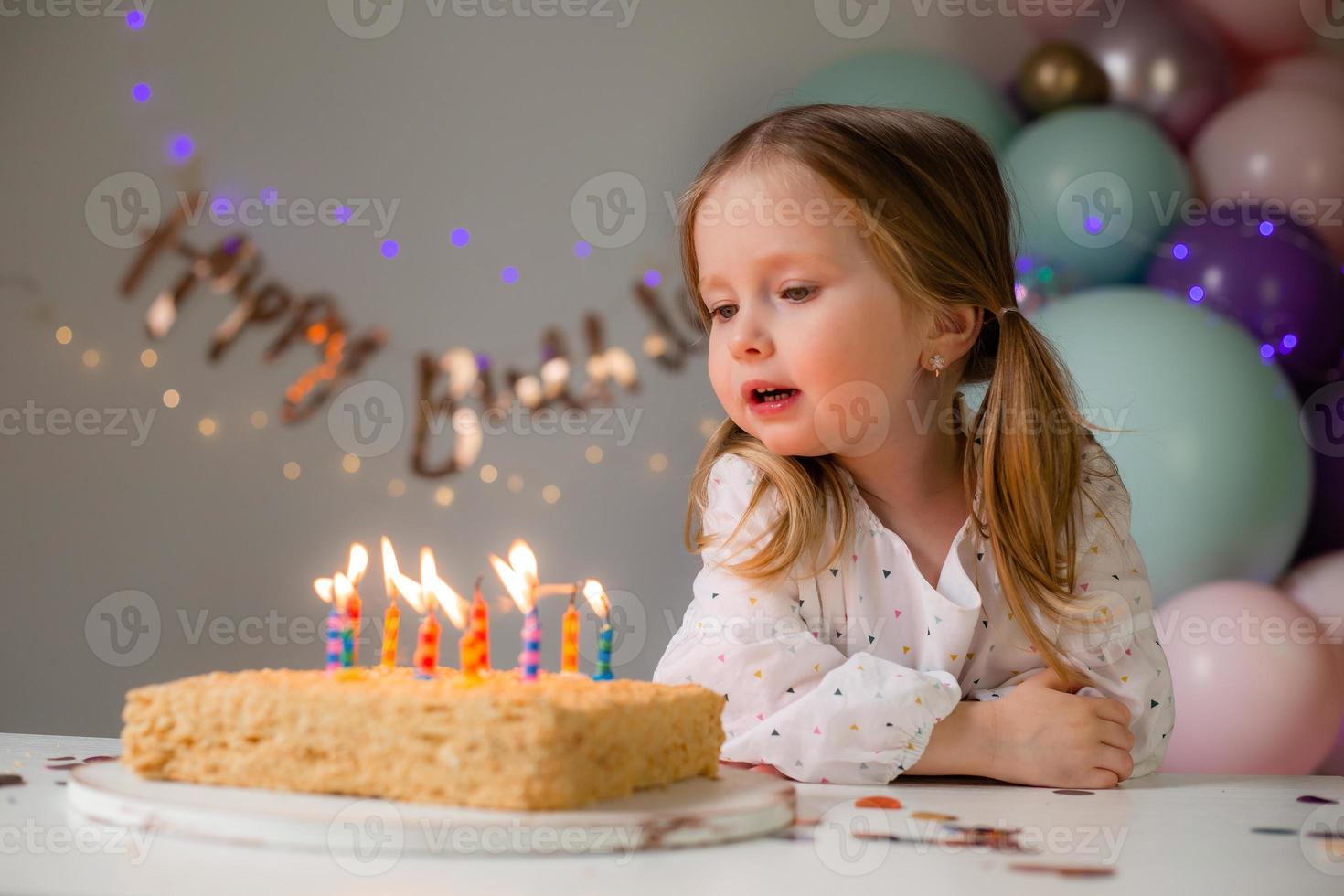 fofa pequeno menina golpes Fora velas em uma aniversário bolo às casa contra uma pano de fundo do balões. criança aniversário foto