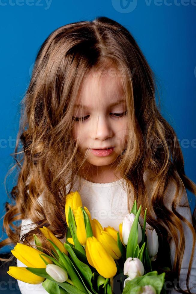 retrato do uma encantador sorridente pequeno menina com uma ramalhete do tulipas dentro dela mãos. estilo de vida. fresco flores internacional mulheres dia. espaço para texto. Alto qualidade foto