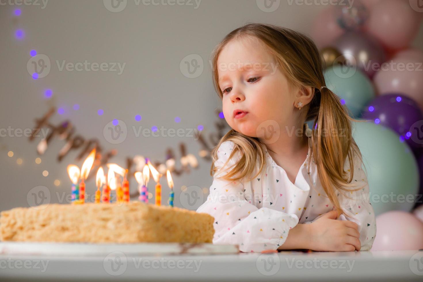 fofa pequeno menina golpes Fora velas em uma aniversário bolo às casa contra uma pano de fundo do balões. criança aniversário foto