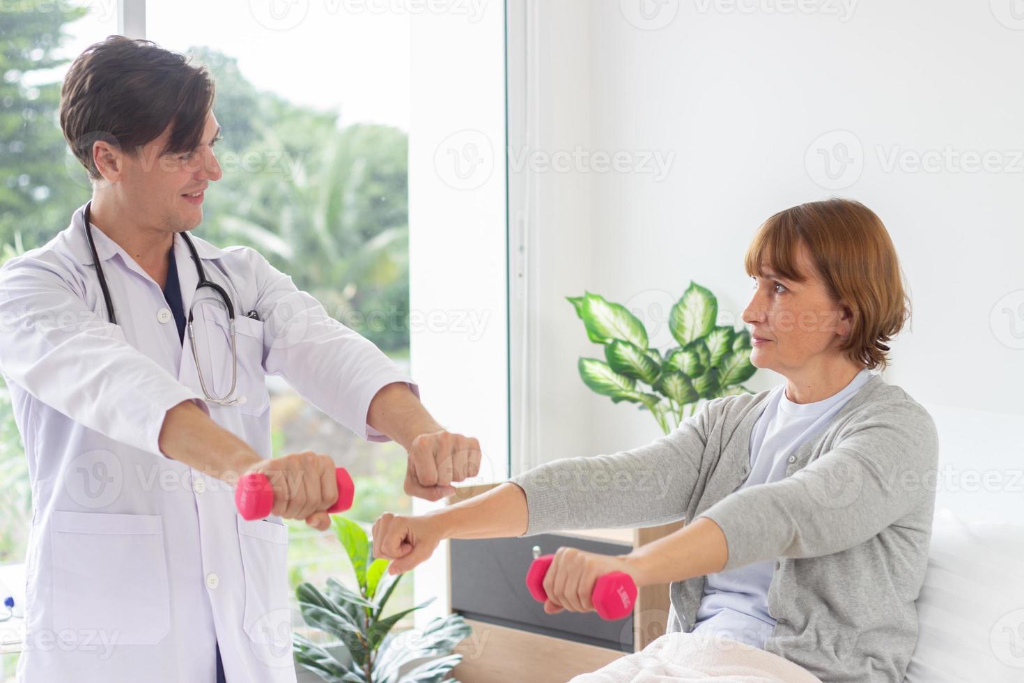 fêmea paciente exercício com halteres dentro a hospital. fisioterapeuta ajudando Senior mulher para lift halteres. a médico é examinando a paciente dentro a hospital foto