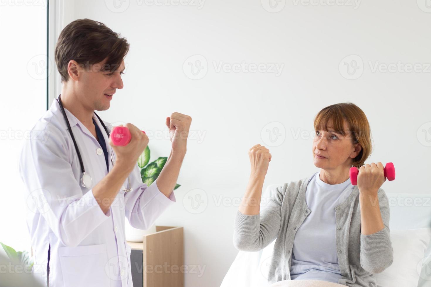 fêmea paciente exercício com halteres dentro a hospital. fisioterapeuta ajudando Senior mulher para lift halteres. a médico é examinando a paciente dentro a hospital foto