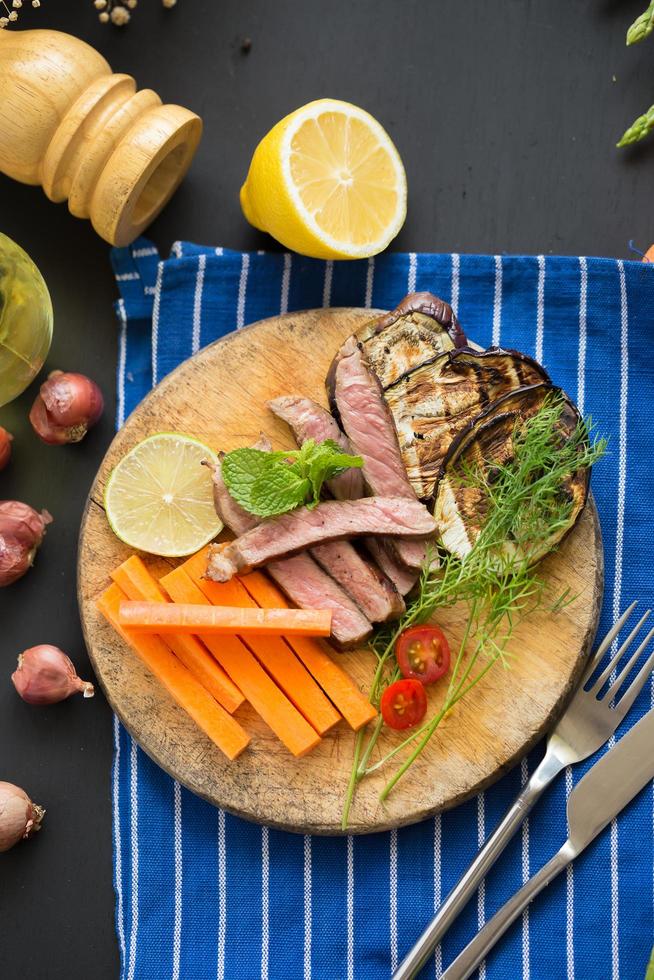 bife grelhado, cogumelos e guarnições na tábua de madeira com cenoura, tomate e limão na toalha de mesa azul na mesa de madeira escura foto