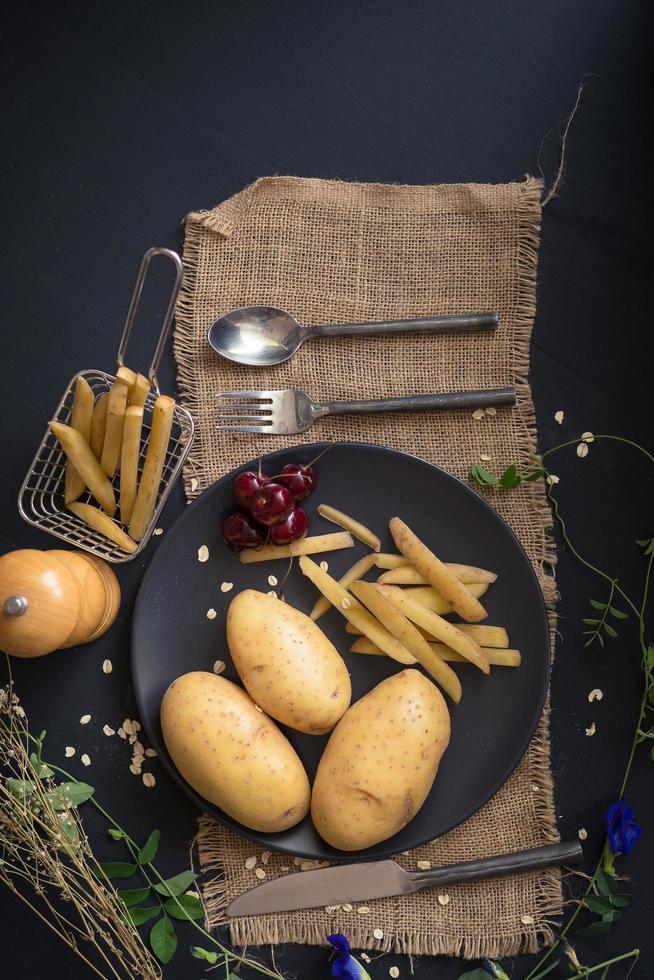 batatas, batatas fritas e cerejas em uma placa preta com utensílios em uma estopa e uma mesa de madeira escura foto