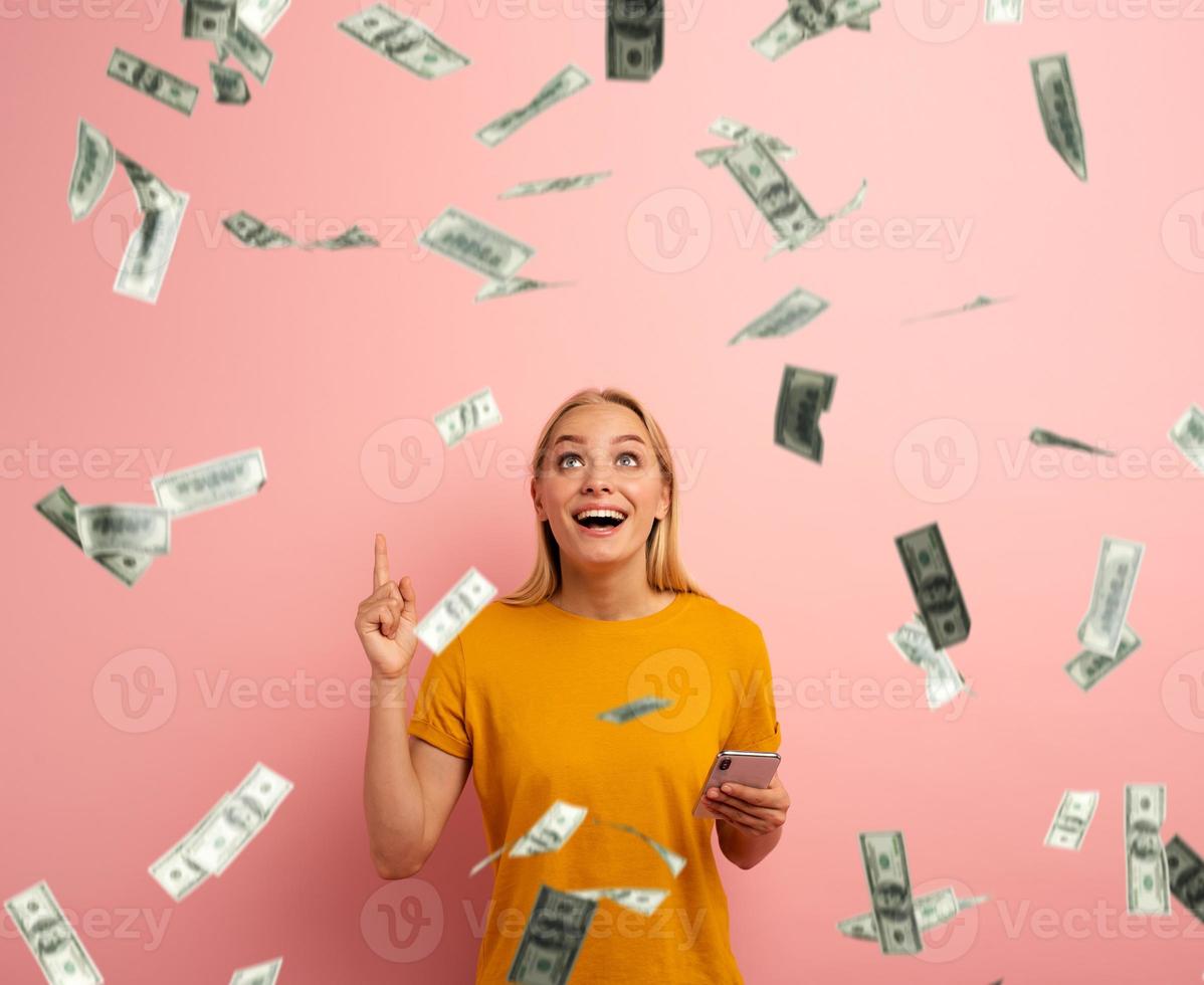 Loiras fofa menina vitórias dinheiro com Smartphone aplicativo. espantado e surpreso expressão face. Rosa fundo foto