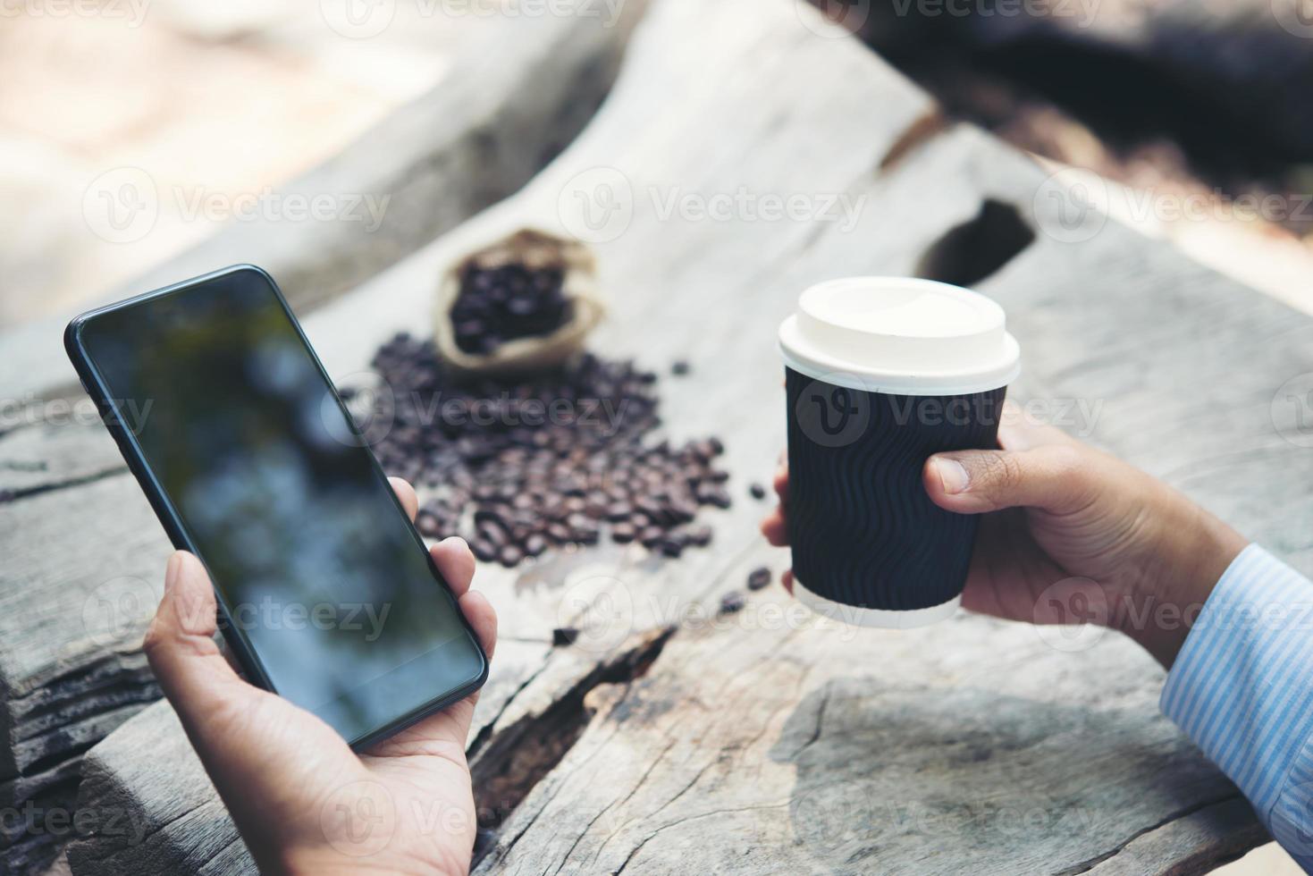 mão do homem segurando uma xícara de café de papel com smartphone sentado à mesa de madeira foto