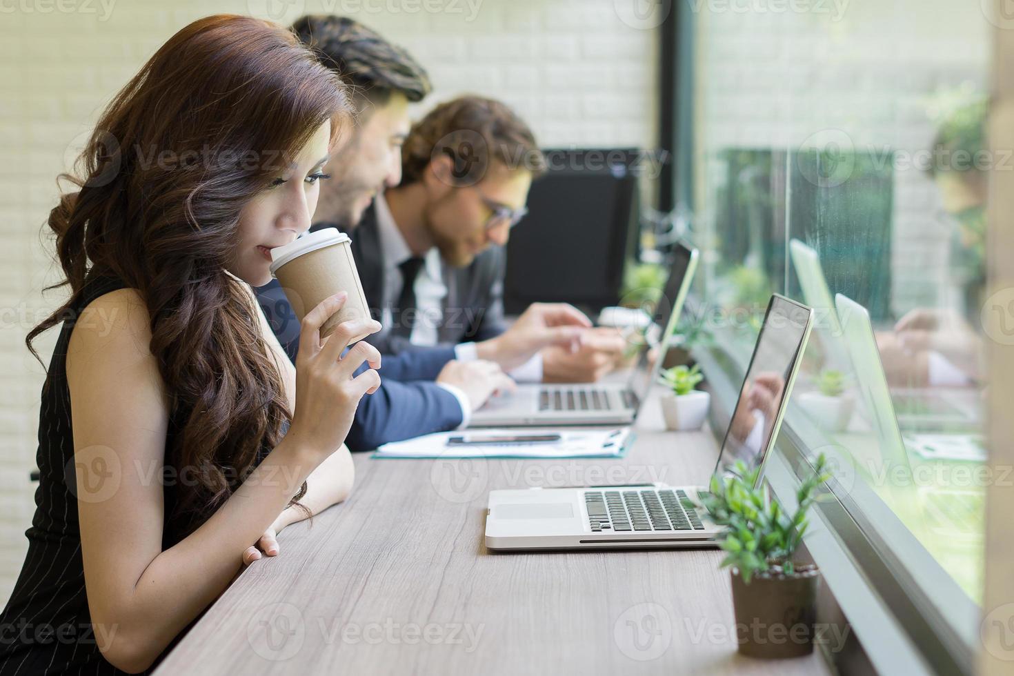 empresária bebendo café olhando para seu laptop enquanto colegas de trabalho interagem em segundo plano foto