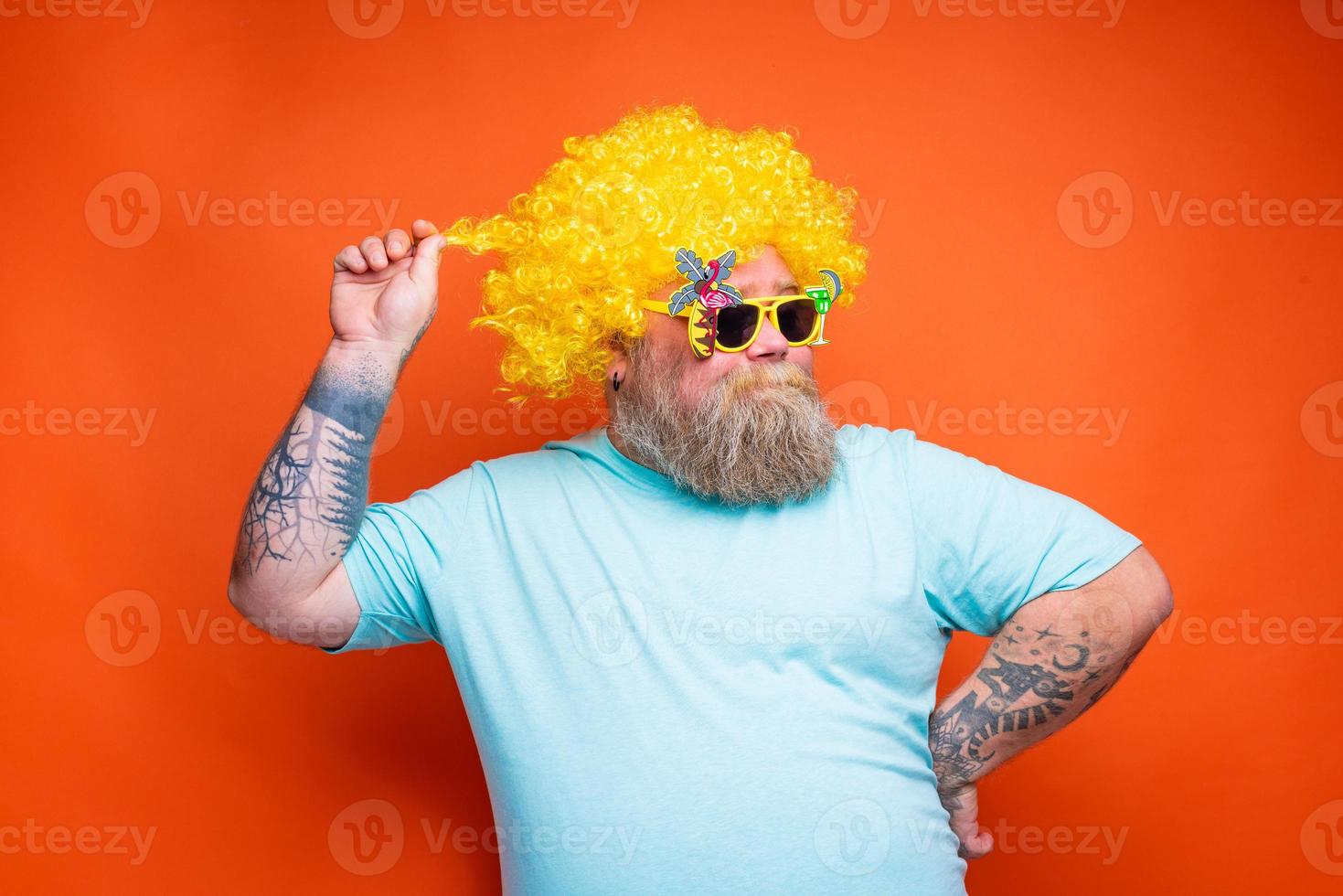 gordo feliz homem com barba, tatuagens e oculos de sol tem Diversão com a amarelo peruca foto