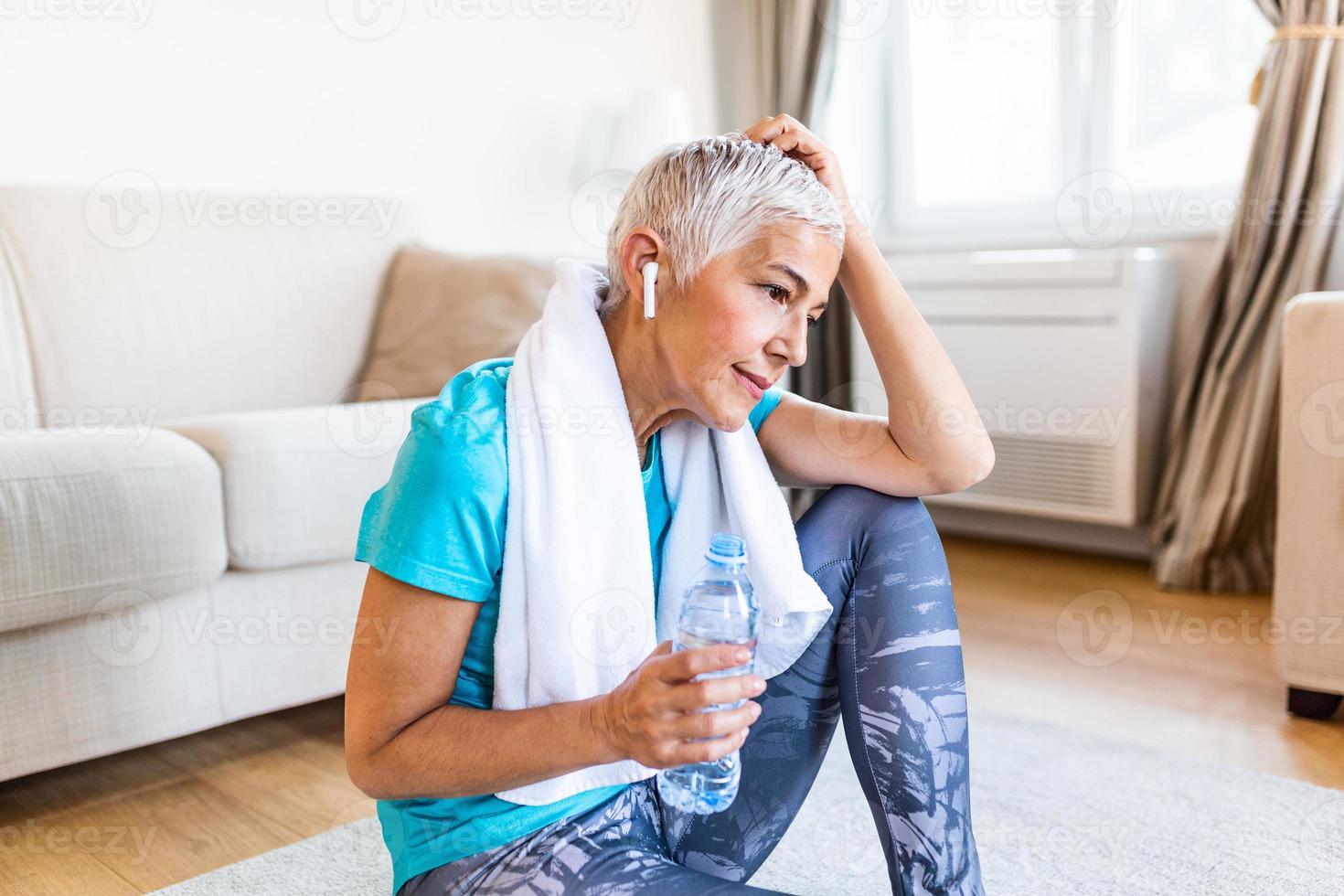 Senior mulher segurando plástico garrafa do água, limpando suor com uma toalha, Exausta depois de a diariamente treinamento. idosos mulher levando uma pausa enquanto exercício às lar. sentado em ginástica esteira e em repouso foto