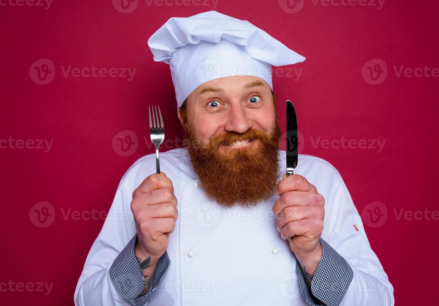 feliz chefe de cozinha com barba e vermelho avental detém talheres dentro mão foto