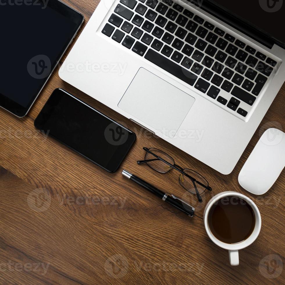 vista superior da área de trabalho com laptop, smartphone, tablet, xícara de café, óculos e caneta na mesa de madeira foto