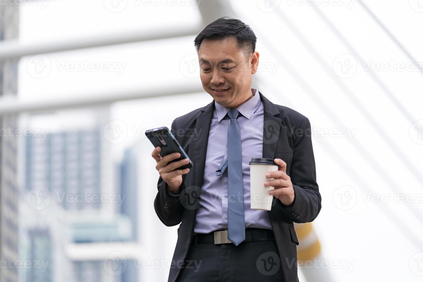 empresário asiático em pé e segurando um telefone celular com prédios de escritórios comerciais ao fundo da cidade foto