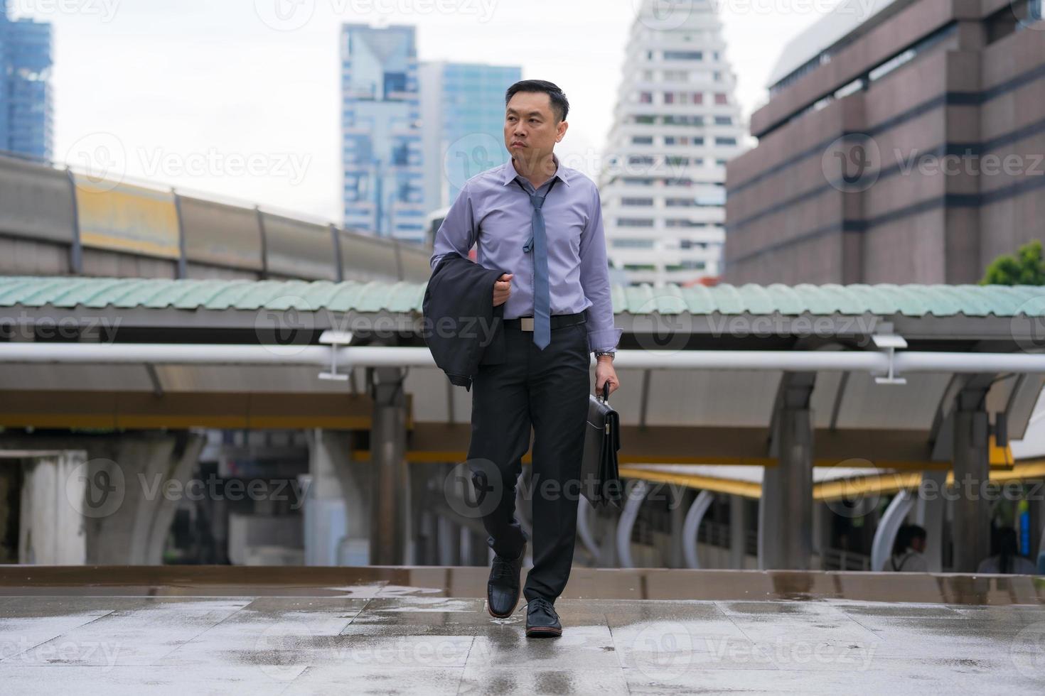 empresário asiático andando e segurando uma pasta com prédios de escritórios comerciais ao fundo da cidade foto