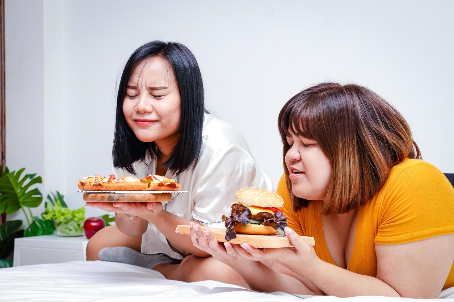 dois gordo meninas comer pizza e hambúrgueres dentro cama dentro a quarto. feliz. a conceito do comendo e saúde Cuidado. foto