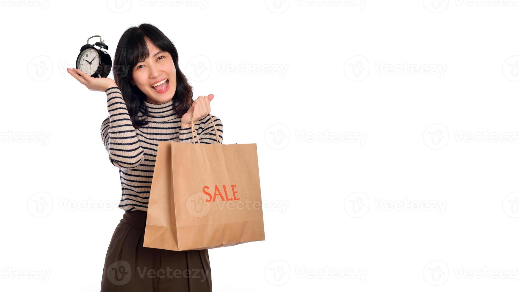 retrato do feliz jovem ásia mulher com suéter camisa segurando alarme relógio e compras papel costas isolado em branco fundo foto