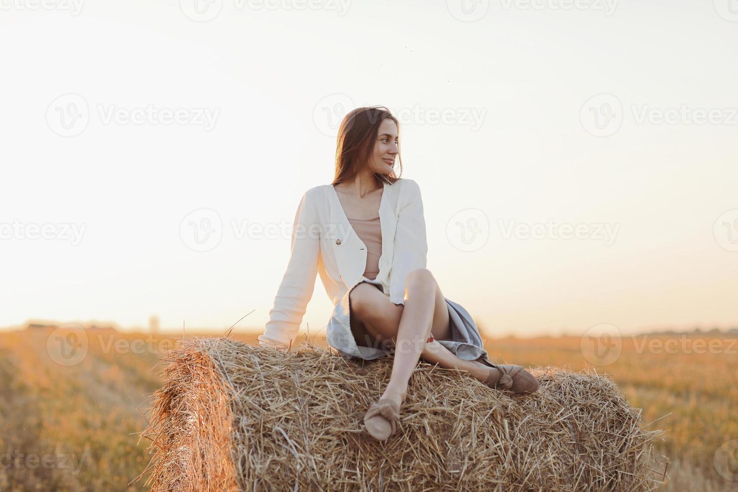 jovem mulher com cabelo comprido, vestindo saia jeans, camisa leve e saco de palha na mão, sentado no fardo no campo no verão. retrato feminino na cena rural natural. conceito de ecoturismo ambiental. foto