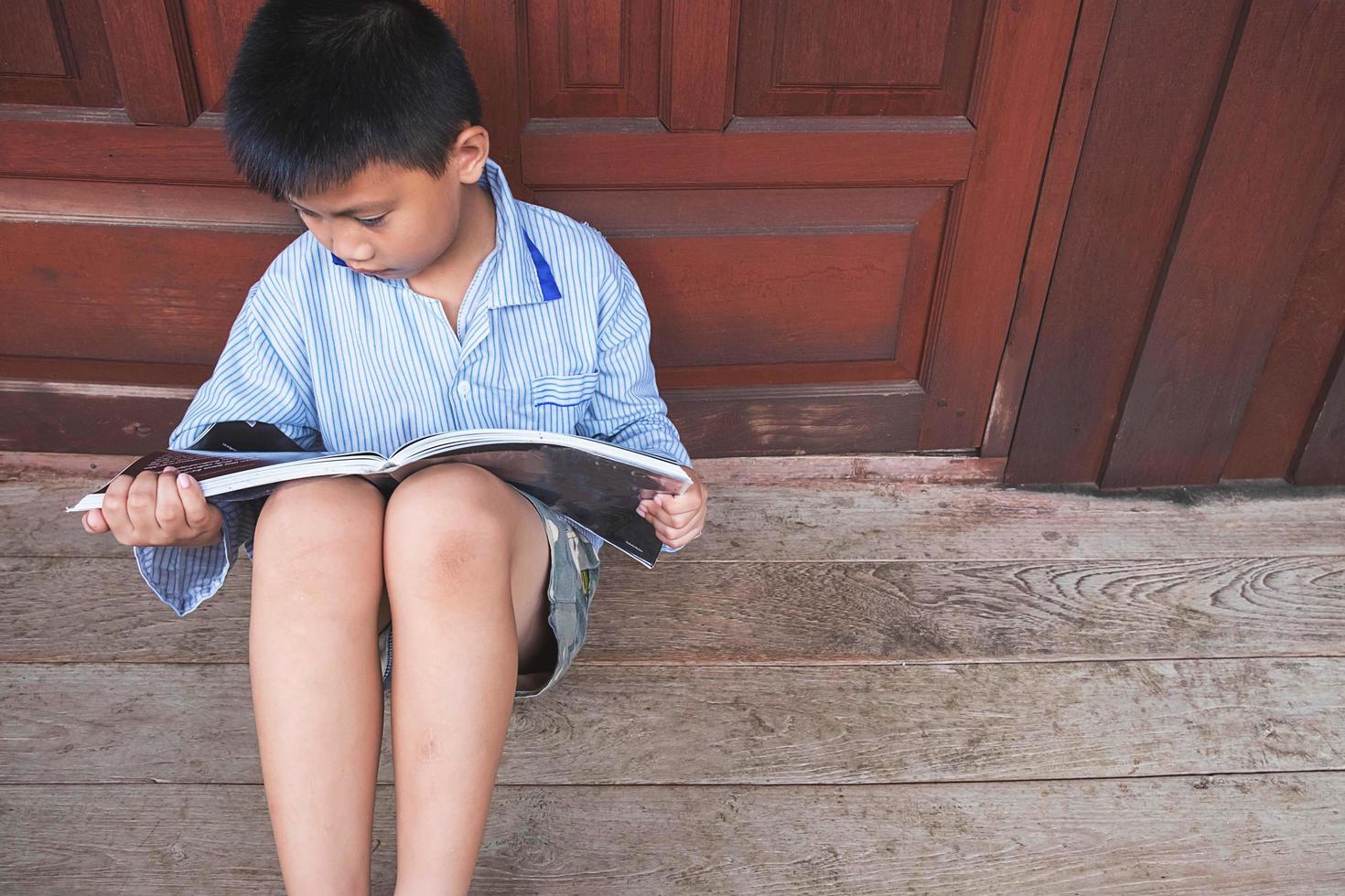 menino sentado contra a porta de madeira lendo um livro no chão de madeira foto