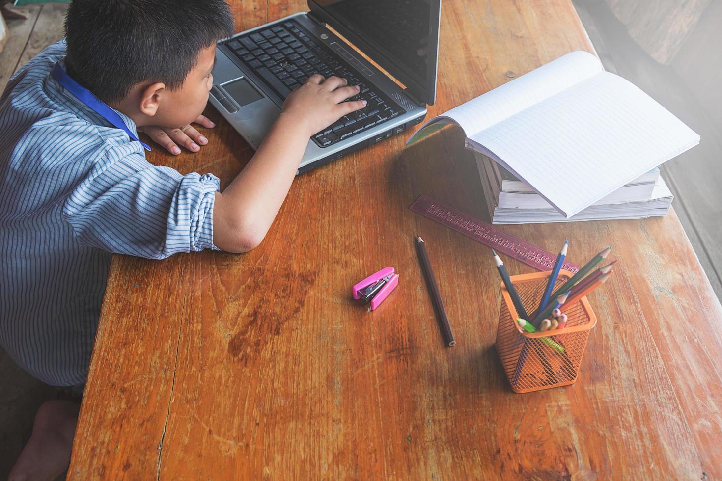 menino trabalhando em um laptop ao lado de um copo de lápis sobre uma mesa de madeira foto