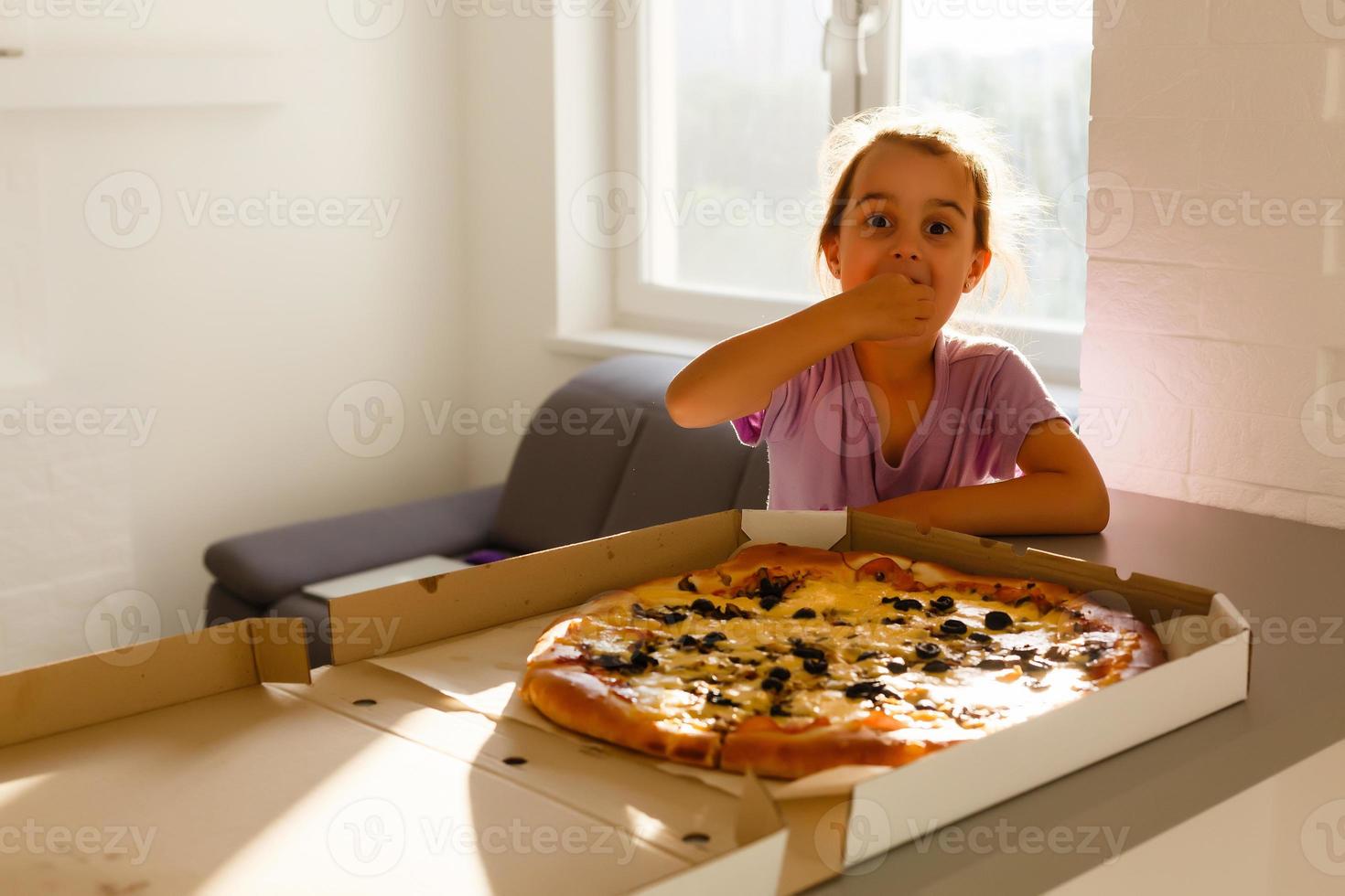 encantador feliz jovem menina rir e morder fora grande fatia do fresco fez pizza. ela sentar às branco cadeira dentro provence estilo interior, sorrir e apreciar ensolarado dia e gostoso refeição. ela tem grandes Loiras cabelo. foto