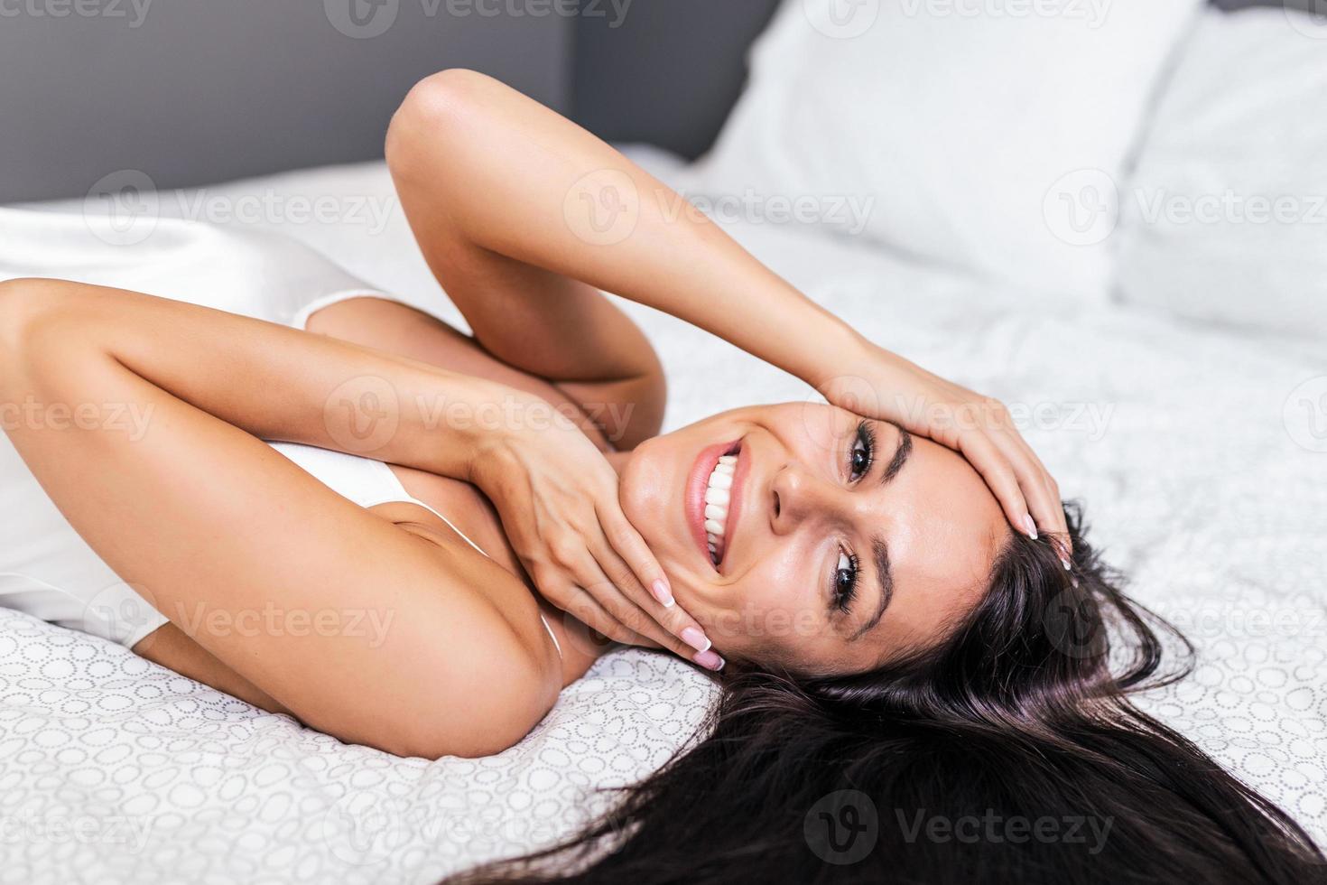 lindo mulher deitado em cama topo visualizar. feliz morena com saudável perfeito cabelo sorridente às Câmera. beleza, alegria, saúde Cuidado, moda conceito foto