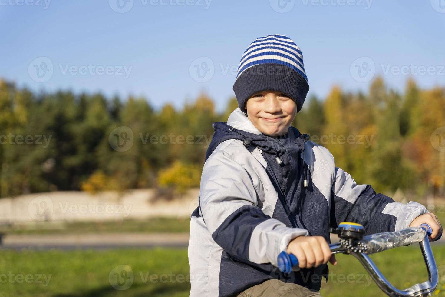 uma alegre criança de cinco anos Garoto passeios uma bicicleta dentro a outono chapéu e Jaqueta contra a fundo do outono árvores foto