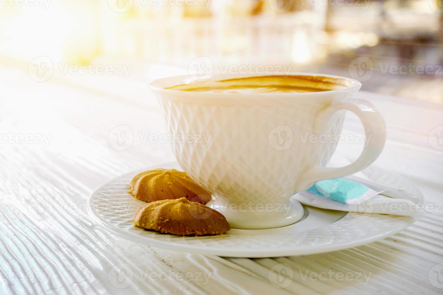 quente café dentro uma branco café copo com biscoitos em branco de madeira mesa em Sol flare fundo. foto