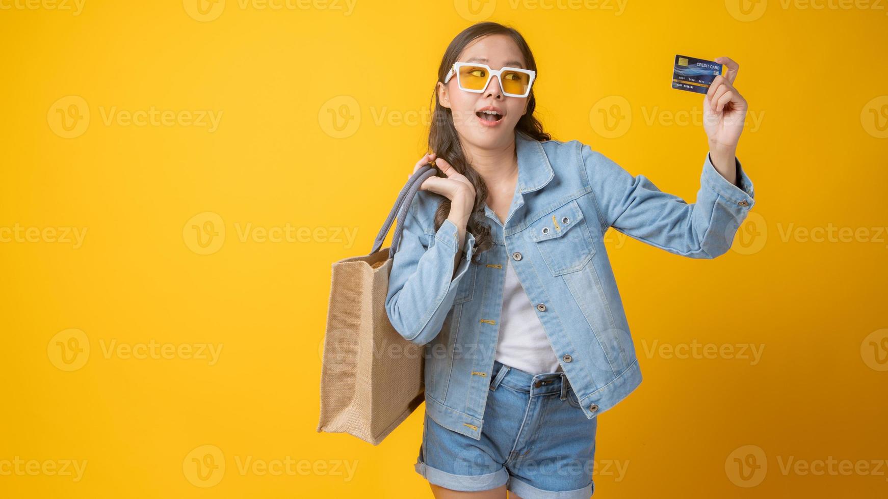 mulher asiática sorrindo e segurando um cartão de crédito e uma sacola de papel no fundo amarelo foto