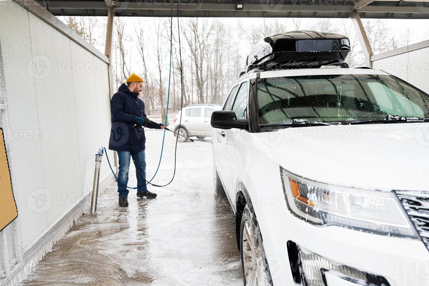 homem lavando carro suv americano de água de alta pressão com rack de teto na lavagem de autoatendimento em clima frio. foto