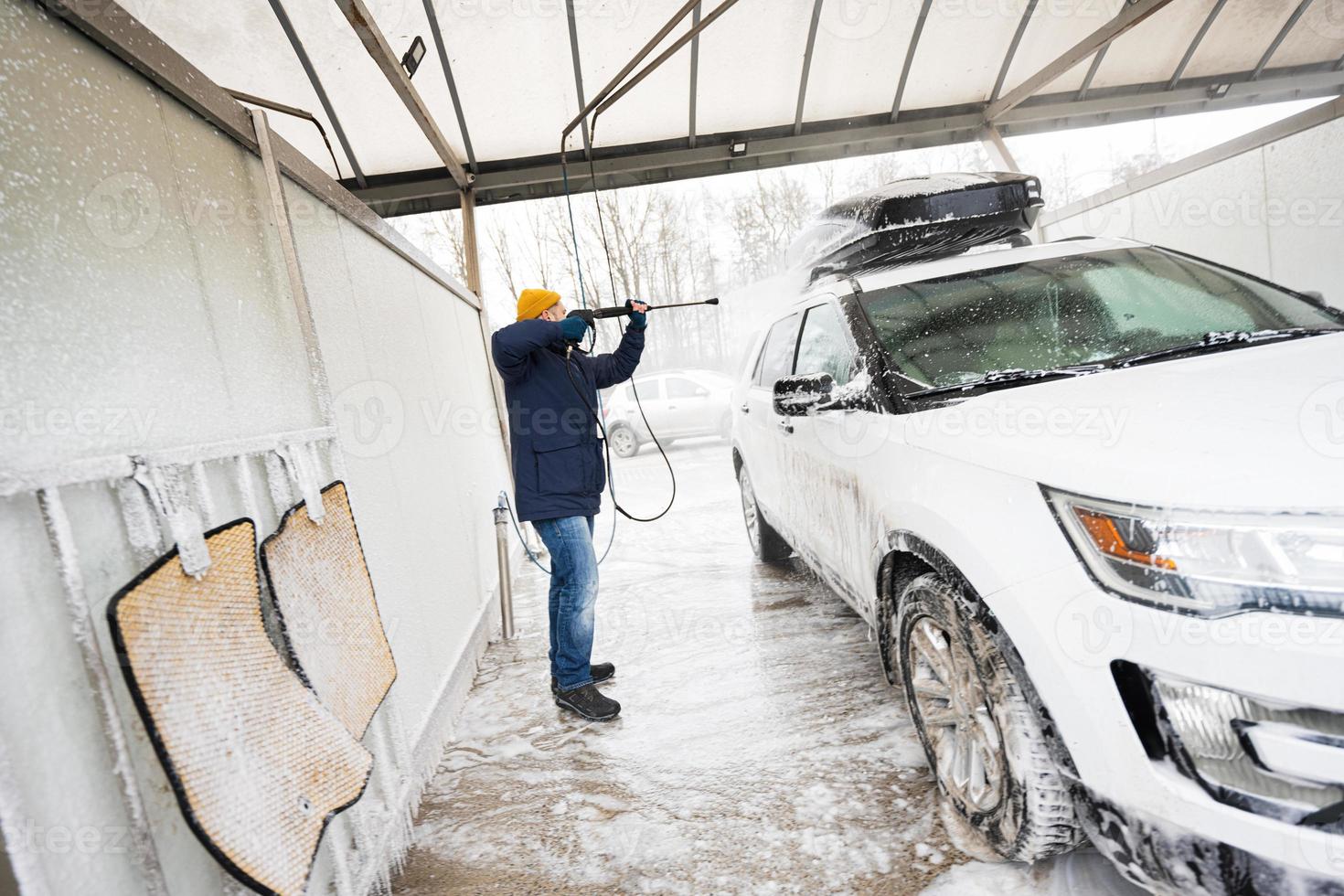 homem lavando carro suv americano de água de alta pressão com rack de teto na lavagem de autoatendimento em clima frio. foto