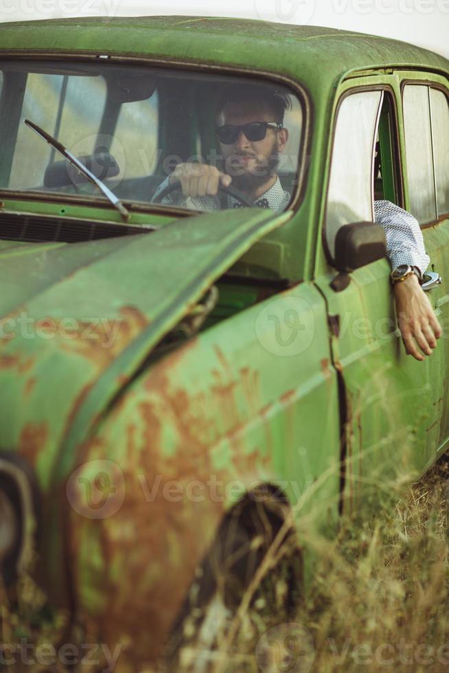 jovem à moda bonito homem, vestindo camisa e oculos de sol, dirigindo velho carro foto