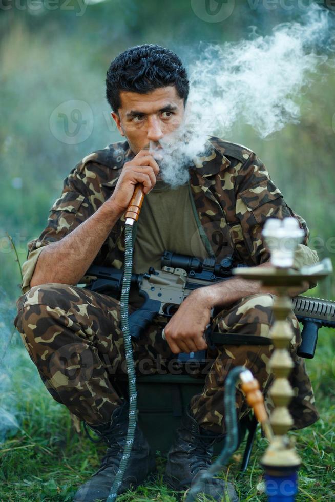 homem dentro camuflar com uma espingarda fuma uma narguilé ao ar livre foto