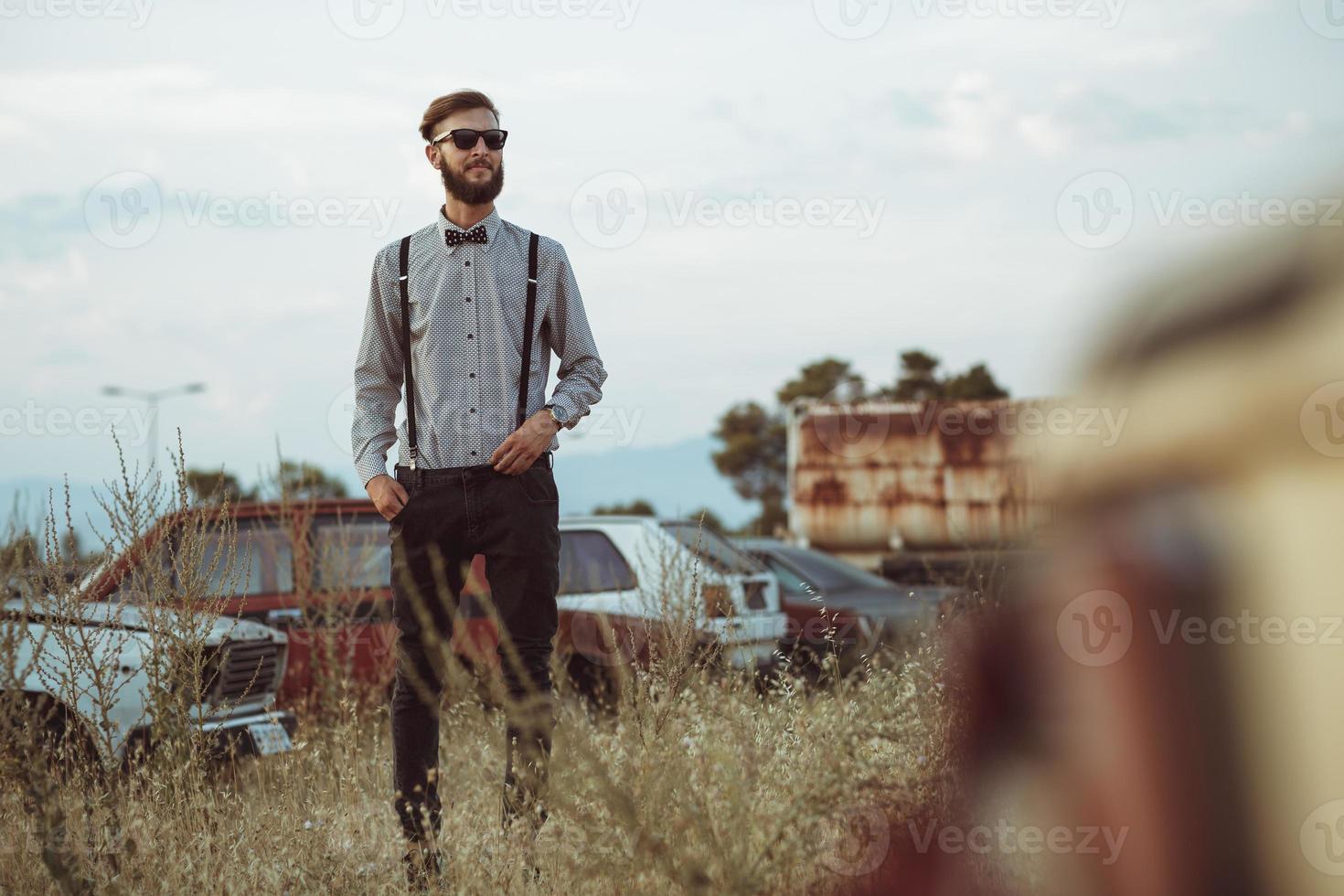 jovem bonito à moda homem, vestindo camisa e gravata-borboleta em a campo do velho carros foto