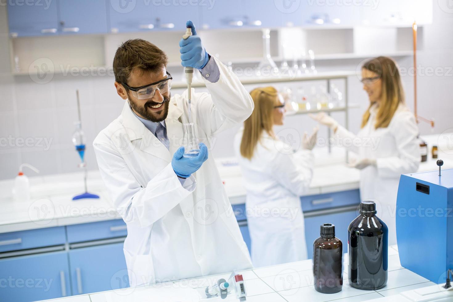 jovens pesquisadores em roupas de trabalho de proteção em pé no laboratório e analisando frasco com líquido foto