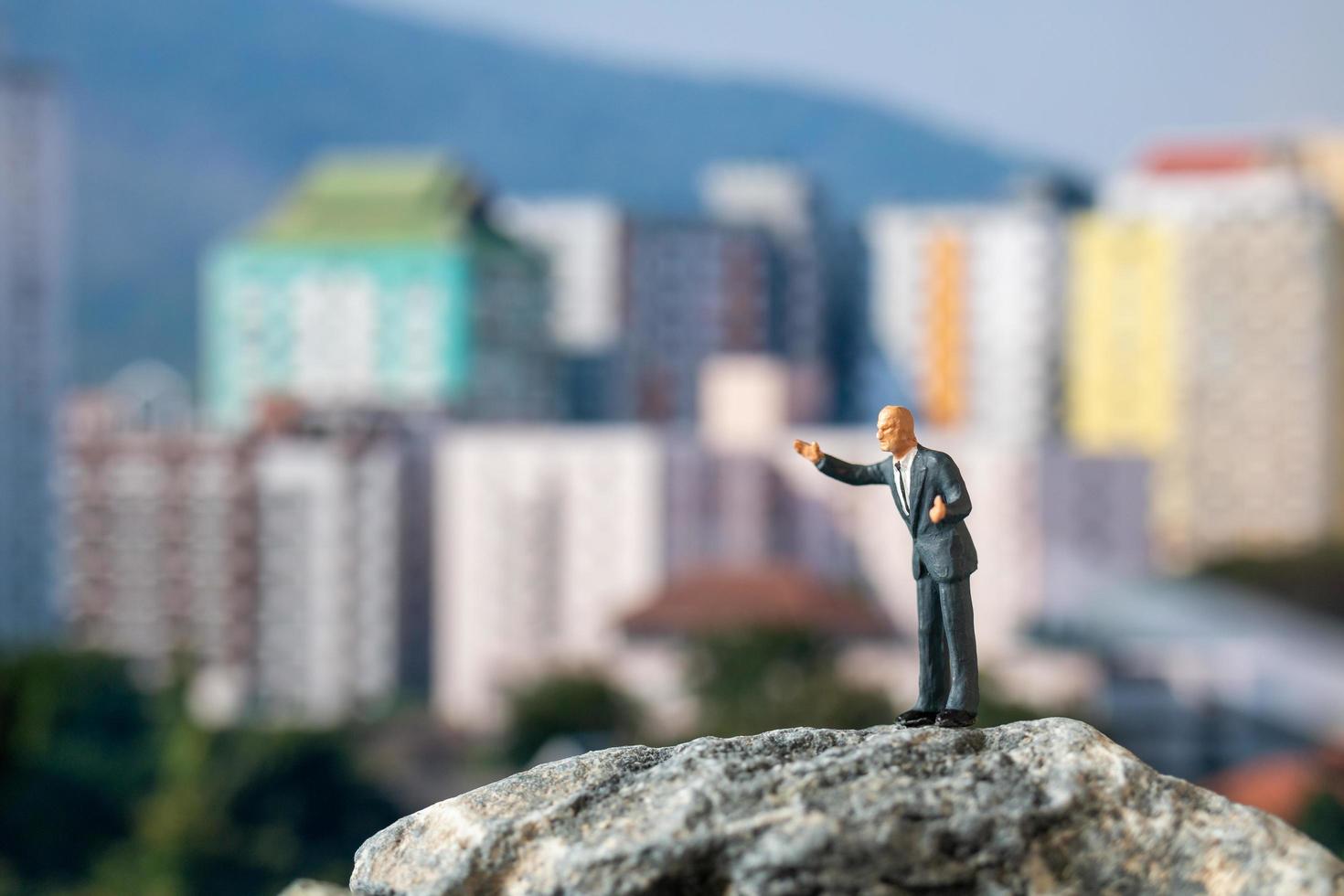 empresário em miniatura de pé em uma rocha com edifícios ao fundo foto