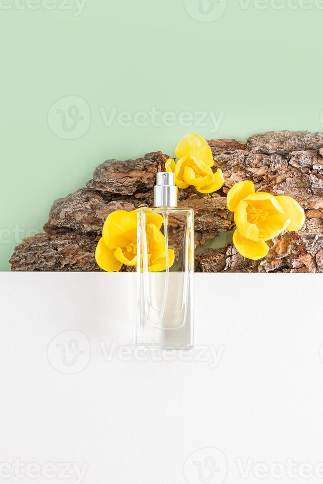 uma transparente garrafa do Cosmético spray mentiras em partes do uma branco texturizado pódio e latido do uma árvore com amarelo tulipas. vertical visualizar. cópia de espaço. foto