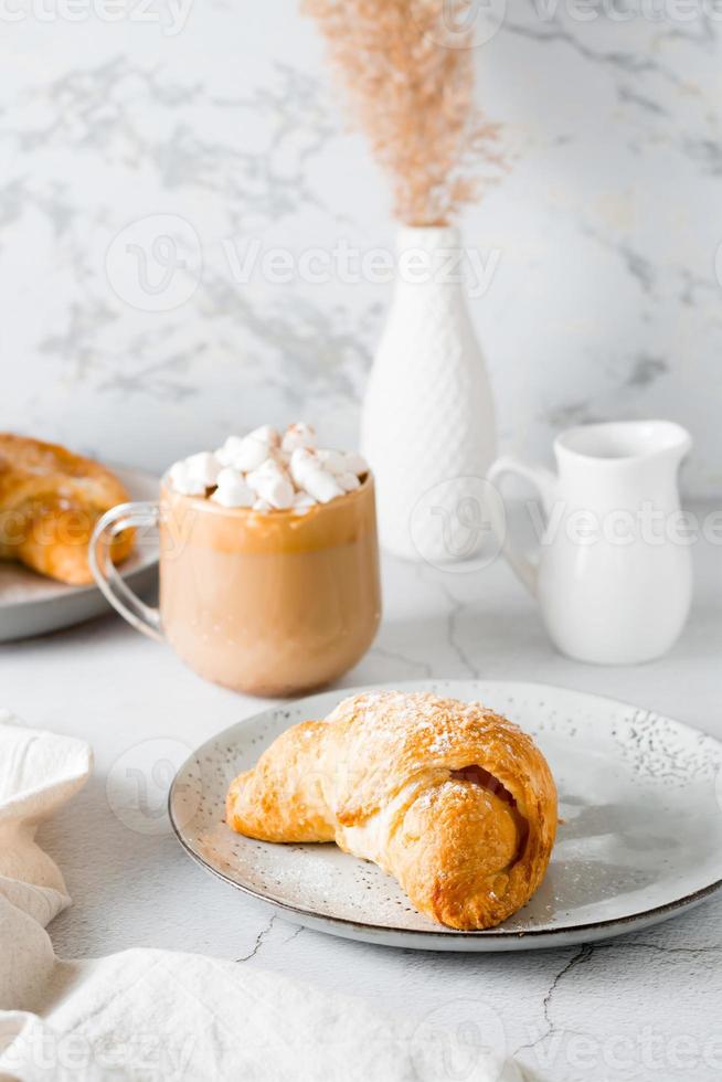 croissant em uma prato e delicioso café com marshmallows dentro uma copo em a mesa. estilo de vida caseiro lanche. vertical Visão foto