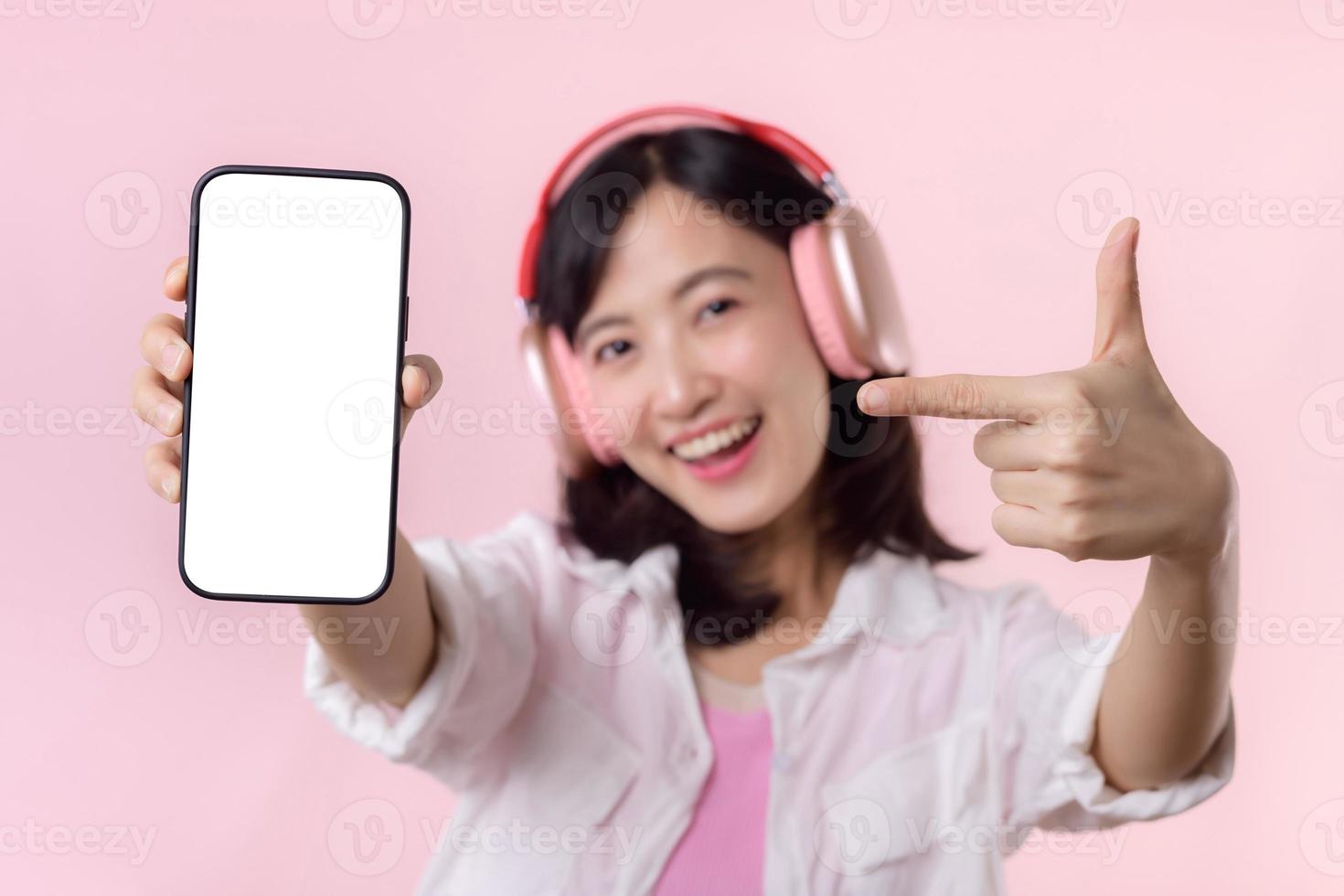 feliz alegre sorridente ásia mulher com sem fio fones de ouvido mostrando em branco tela Móvel telefone ou Novo Smartphone música inscrição propaganda brincar isolado em Rosa estúdio fundo. foto