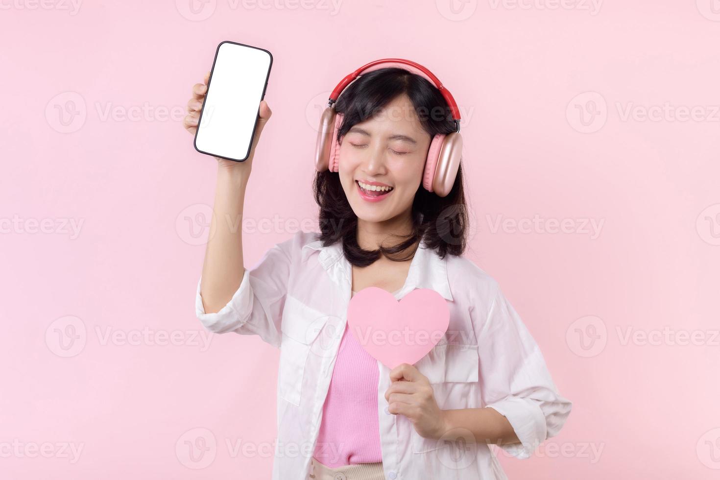 feliz alegre sorridente ásia mulher com mão segurando coração e fones de ouvido mostrando em branco tela Móvel telefone ou Novo Smartphone música inscrição propaganda brincar isolado em Rosa estúdio fundo. foto