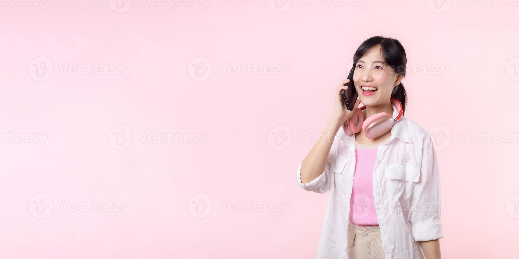 retrato jovem atraente ásia mulher feliz sorrir usando Smartphone com fone de ouvido, fone de ouvido isolado em Rosa estúdio fundo. bonita fêmea pessoa usando Móvel telefone. música conectados estilo de vida conceito. foto