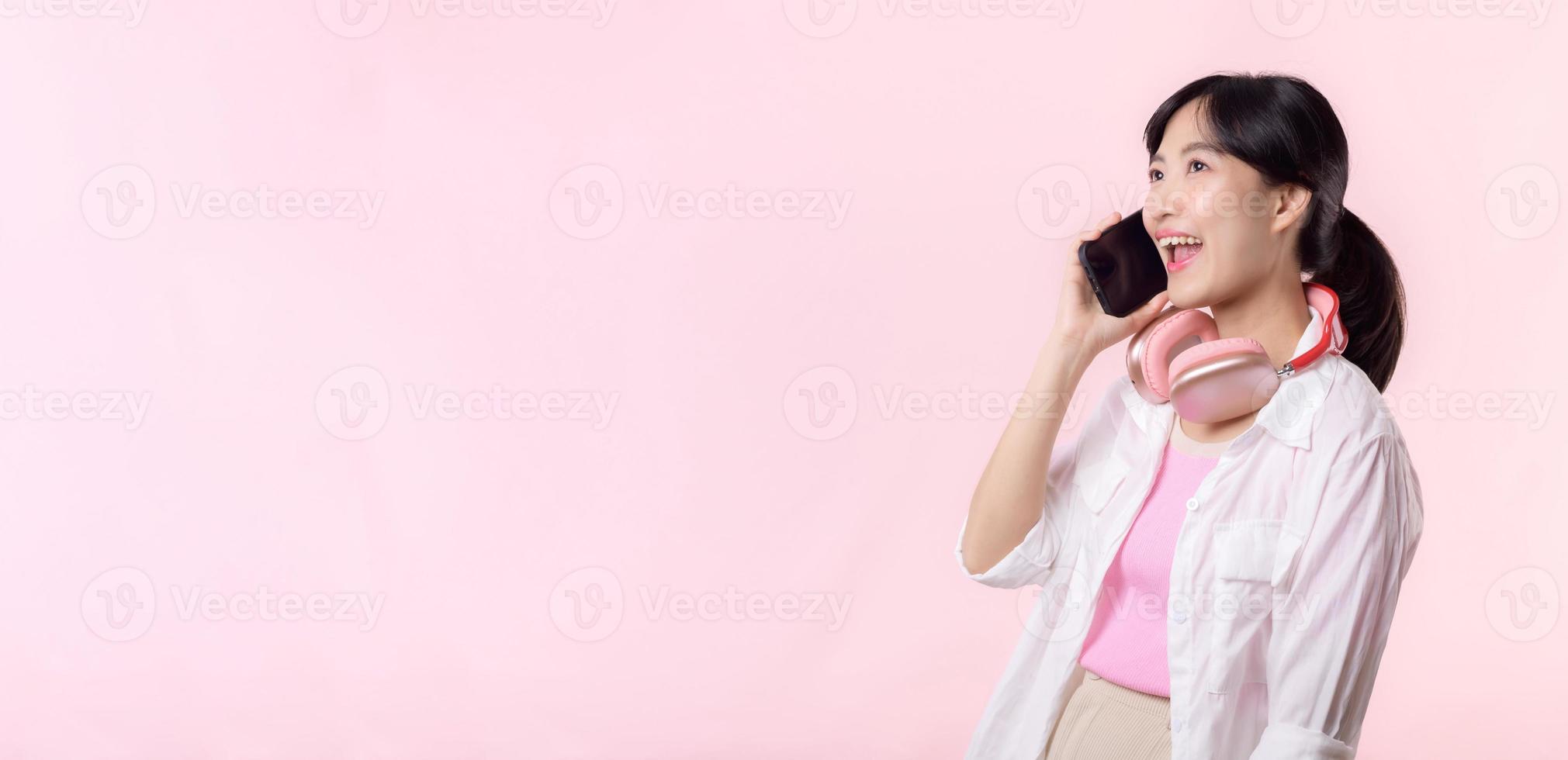 retrato jovem atraente ásia mulher feliz sorrir usando Smartphone com fone de ouvido, fone de ouvido isolado em Rosa estúdio fundo. bonita fêmea pessoa usando Móvel telefone. música conectados estilo de vida conceito. foto